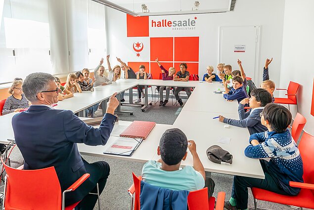 Die Kinder sitzen an Tischen im Rechteck und heben die Finger für Fragen.