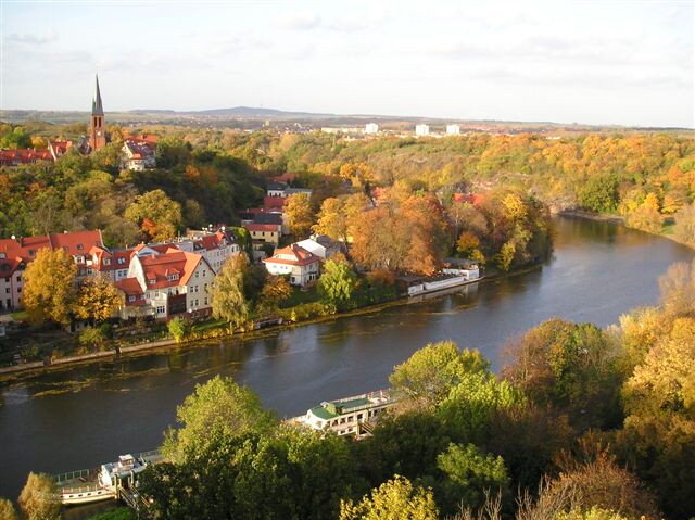 Herbstlandschaft rechts und links eines Flusses, Schiffsanleger, kleine Häuser und hohe Kirche 