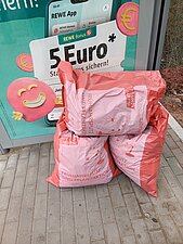 Drei befüllte Müllsäcke stehen vor einem grünen Container.