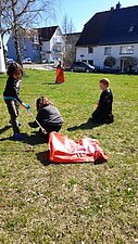 Vier Kinder befinden sich auf einer Grünfläche. Ein Kind hält eine Greifzange in der Hand. Ein anderes Kind hält einen Müllsack.